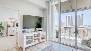 Ein modernes Wohnzimmer mit großem Fernseher, weißen Möbeln und einem Balkon mit Blick auf die Stadt.