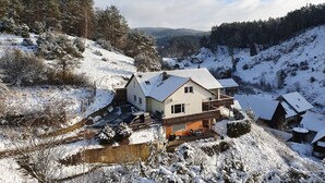 A snow-covered house with a sloped roof, surrounded by trees and hills.