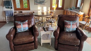 A cozy living room with leather armchairs, a dining table, and a small side table with a laptop and remote controls.