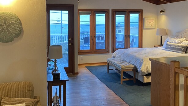 A bedroom with a large bed, a wooden bench, a small table with a lamp, and a view of the sea through large windows.