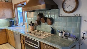 A kitchen with a gas stove, oven, and hanging pots. There is a clock on the wall and a coffee maker on the counter.