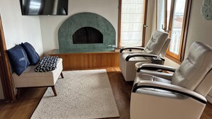 A living room with a fireplace, two leather chairs, a sofa with patterned cushions, a flat-screen TV mounted on the wall, and a large window with blinds.