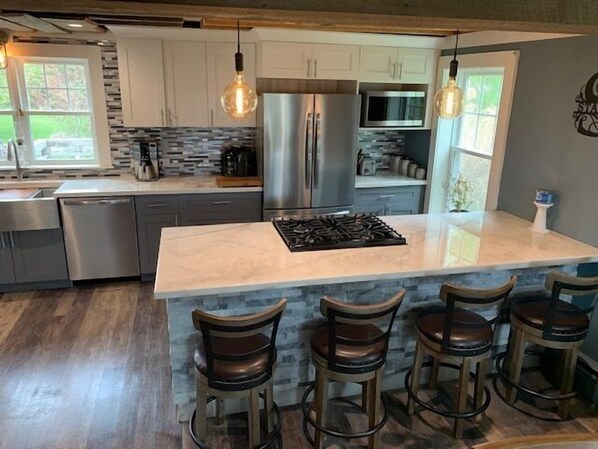 A modern kitchen with a central island, bar stools, stainless steel appliances, and hanging pendant lights.