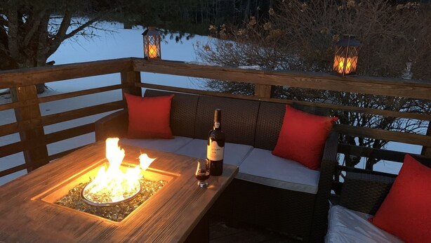 Eine Holzterrasse mit einer Feuerstelle, einer Weinflasche und einem Glas auf einem Tisch. Rote Kissen liegen auf einer Bank und im Hintergrund stehen Laternen.