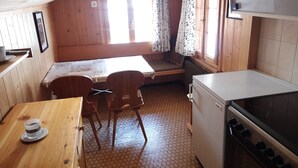 A compact kitchen with wooden walls, a small dining table, two chairs, a refrigerator, and a stove.