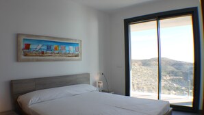 A bedroom with a large bed, a nightstand, a painting of beach chairs, and a view through a large window. - Canyelles Mar Casa 1 (Roses)