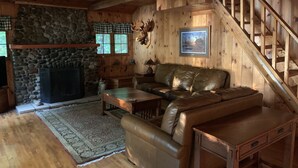 Ein gemütliches Blockhaus-Interieur mit Kamin aus Stein, Ledersofa, Holztreppe und einem gerahmten Bild an der Wand.
