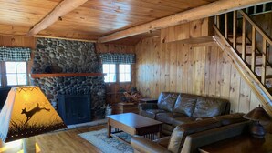 A cozy cabin interior with a stone fireplace, wooden walls, a leather sofa, and a staircase.