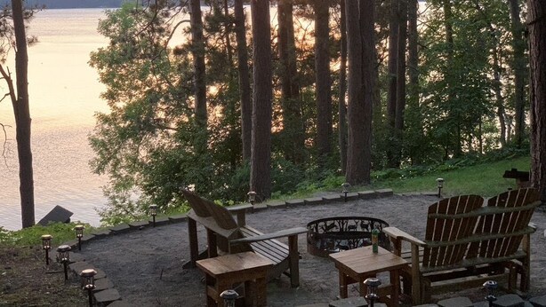 Ein Sitzbereich am Seeufer mit Holzsesseln und Tischen, umgeben von Bäumen und Blick auf den Sonnenuntergang über dem Wasser.