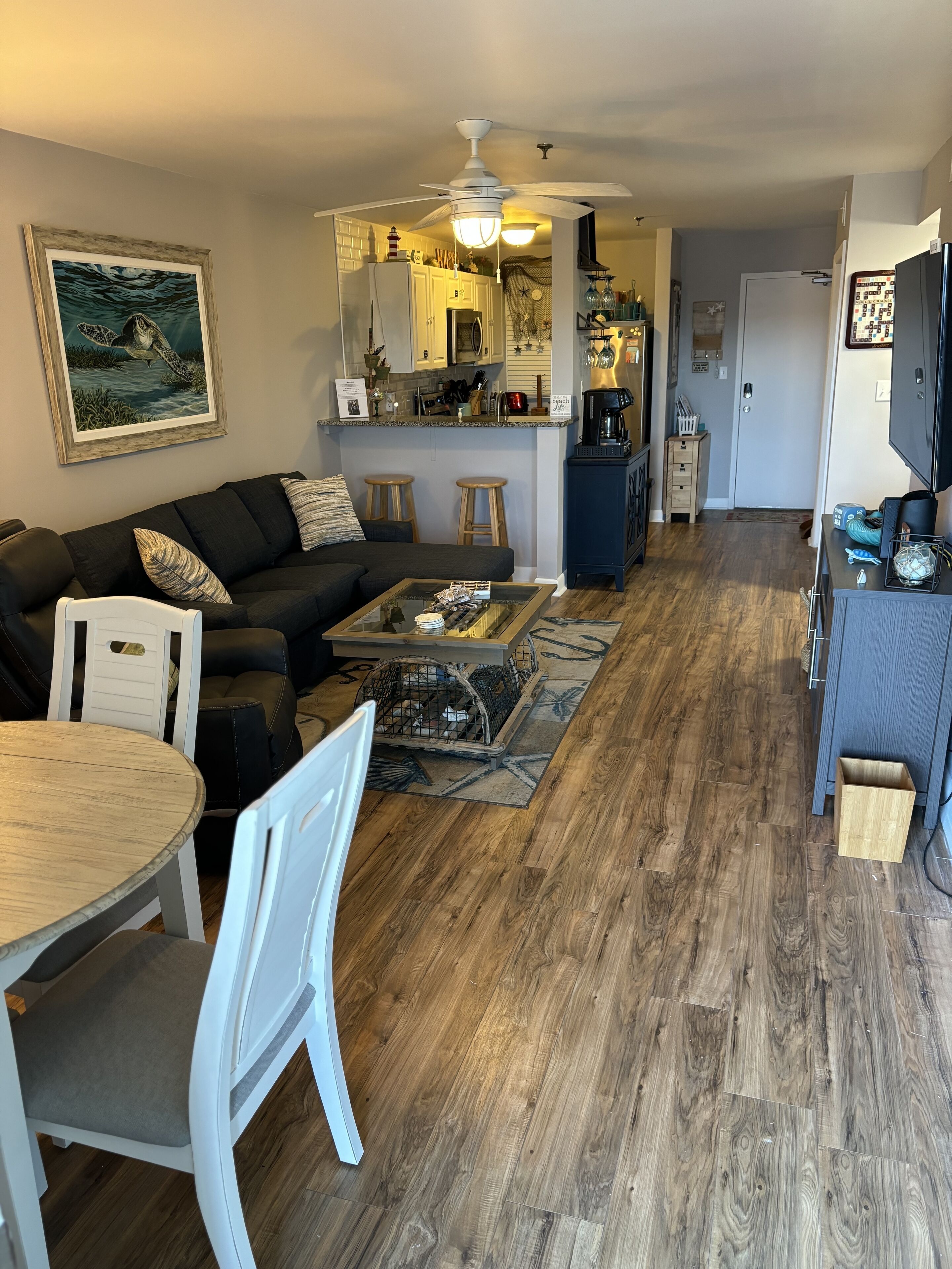 Ein Wohnzimmer mit einem dunklen Sofa, einem Couchtisch und einem Essbereich mit weißem Tisch und Stühlen. Im Hintergrund ist eine Küche mit Deckenventilator und Kühlschrank erkennbar.