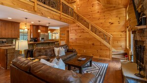 Interior espacioso de una cabaña de troncos, con una sala de estar que incluye un sofá de cuero, una mesa de centro y una chimenea. Una escalera de madera con barandilla conduce a un nivel superior.