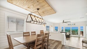 A spacious dining area with a wooden table and chairs, a ceiling fan, and a view of the ocean through large windows. - 🐚 Silver Beach Retreat | Beachfront | FREE VIP Package | Beach Chair Service (Destin)