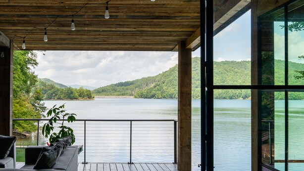 Une terrasse en bois avec un canapé et un pot de fleurs, donnant sur un lac bordé de collines.