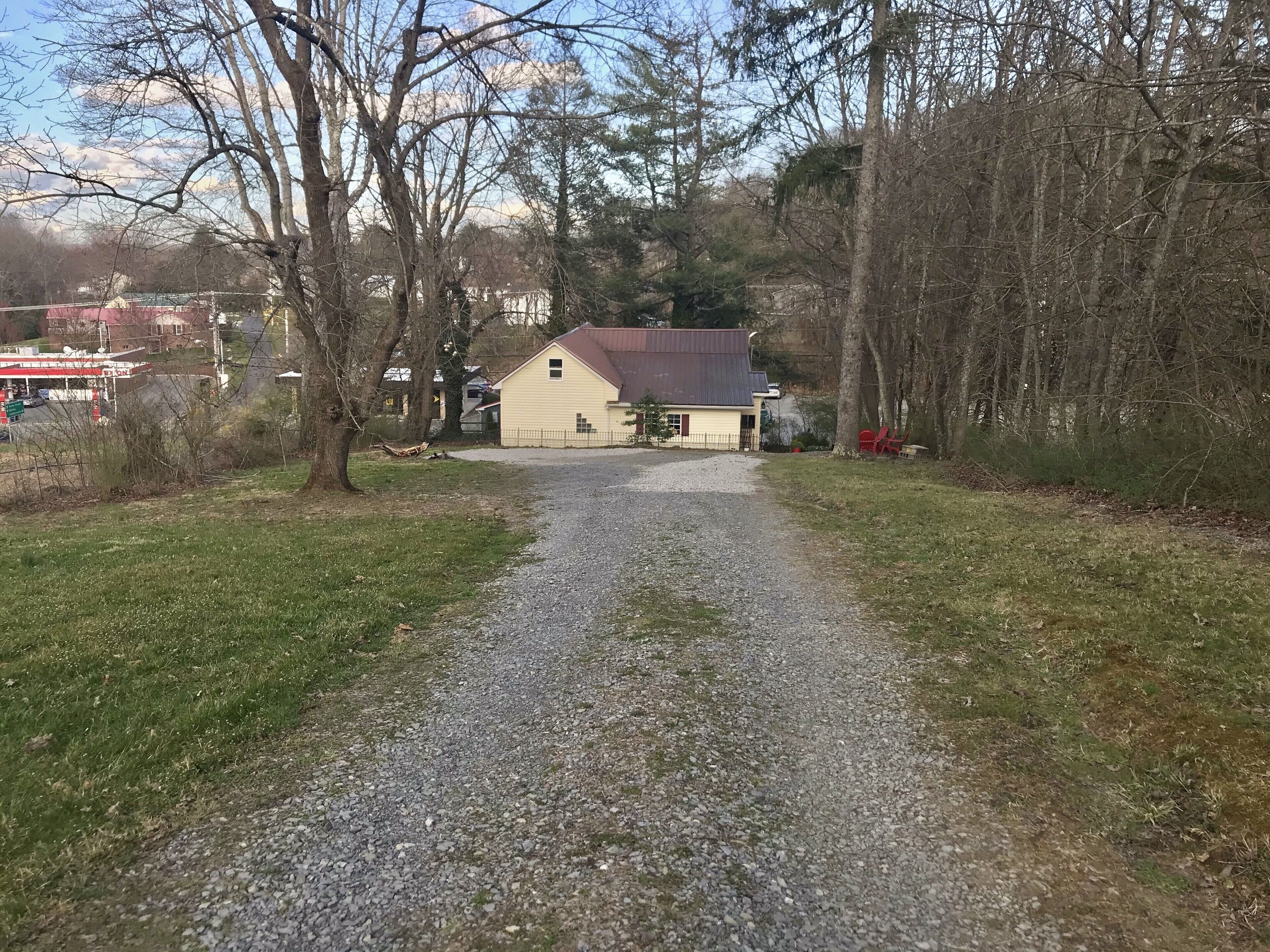 A gravel path leading to a yellow house with a red roof, surrounded by trees and a gas station in the background.
