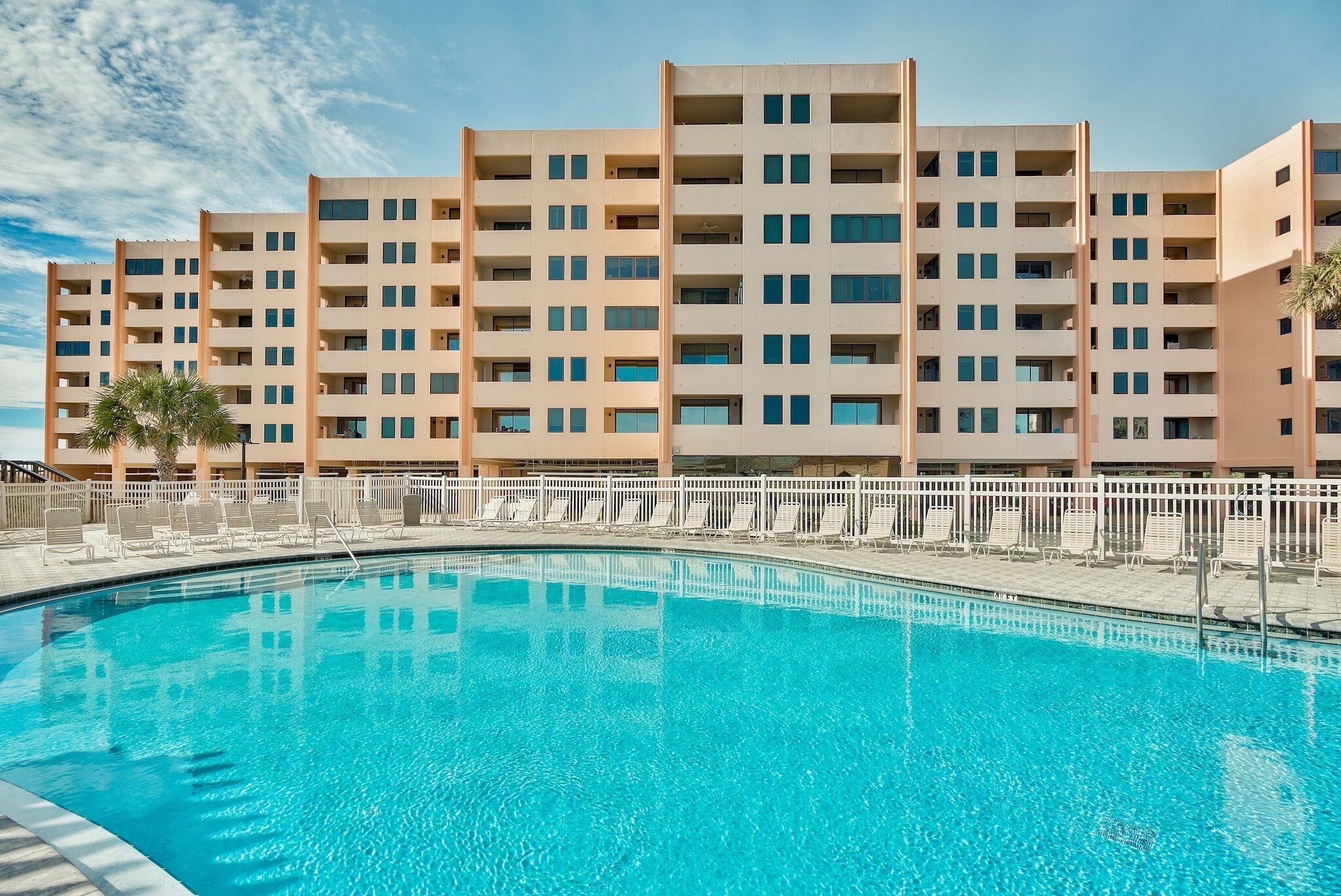 A hotel with a swimming pool, multiple balconies, and a white fence surrounding the pool area.
