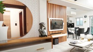 Un soggiorno moderno con TV montata su pannello di parete in legno, uno specchio rotondo e un'area pranzo con tavolo e sedie bianche.