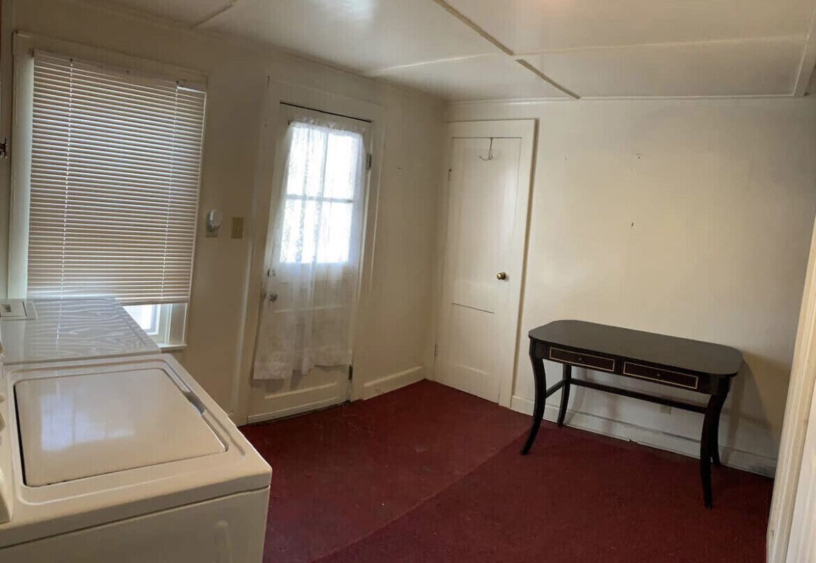 Una habitación con lavadora, una mesa, una puerta y un ventanal con persianas.