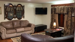 Ein Wohnzimmer mit einem braunen Ledersofa, einem schwarzen Ottomanen, einem Holztisch und einem an der Wand montierten Fernseher.