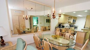 Un coin repas avec une table en verre entourée de chaises en osier, d’un canapé et d’un grand miroir qui reflète la pièce.