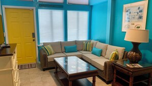 Sala de estar con sofá, mesa de centro y una lámpara. Se observa una puerta amarilla y una ventana con persianas.