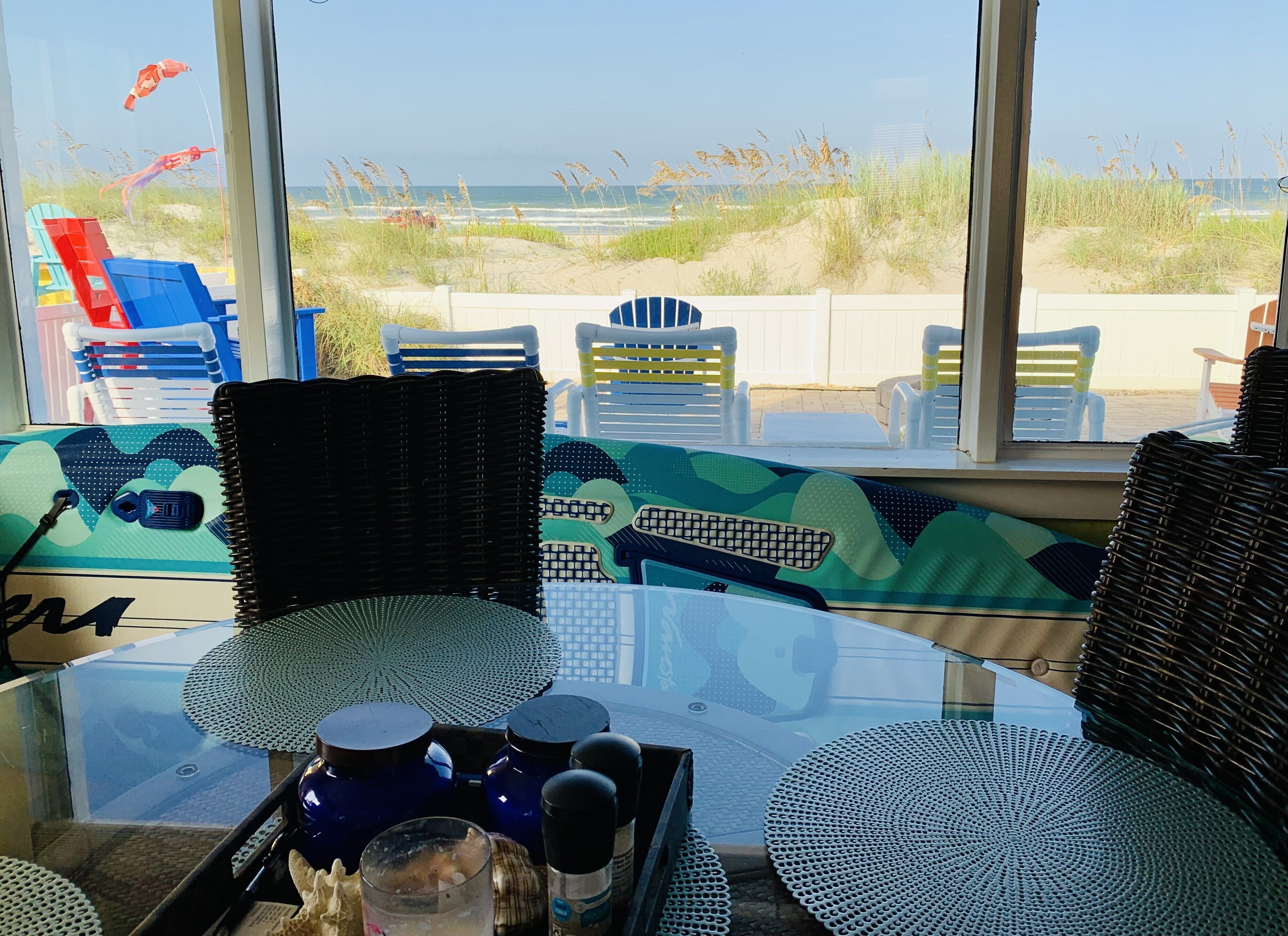 A beachside view from a room with wicker furniture, a glass table, and a view of the beach and dunes.