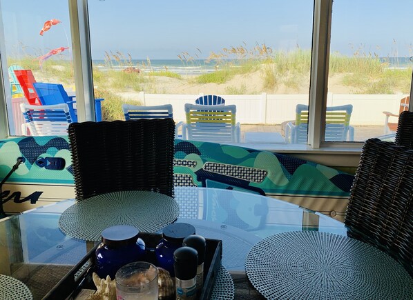 A beachside view from a room with wicker furniture, a glass table, and a view of the beach and dunes.