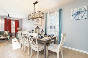 Een eetgedeelte met een houten tafel, witte stoelen en een kroonluchter. Er is een plafondventilator, een raam met gordijnen en een muur met een lijstwerkfoto.