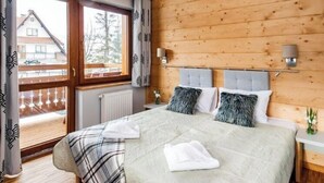 A bedroom with a wooden ceiling, a large bed with grey headboards, a window with curtains, and a view of a building outside. - Lipki Park Resort by Interhome (Zakopane)