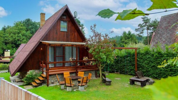 Une cabane en bois avec un toit en pente, une véranda avec des chaises en bois et un jardin délimité par une clôture en bois.