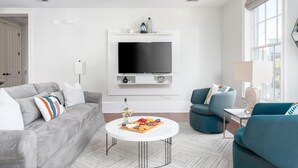 Ett modernt vardagsrum med en grå soffa, en blå fåtölj, ett soffbord med en fruktfat på, en platt-TV som är monterad på en vit vägg och ett stort fönster som släpper in naturligt ljus.