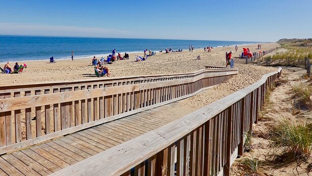 Une promenade en bois mène à une plage de sable où des gens se détendent et prennent des bains de soleil.