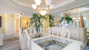 A dining area with a glass-top table, white chairs, a chandelier, and decorative plants. - Elegant Oceanfront 2 Bedroom 2 Bath Ocean View Condo at Maisons-Sur-Mer in Myrtle Beach (Myrtle Beach)