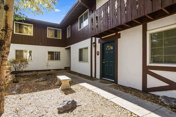 A two-story house with a dark brown exterior and white walls. There is a green door with a decorative design, a stone bench, and a small garden area with rocks and a tree. - Fall River Escape; Perfect Getaway on Fall River! (Estes Park)