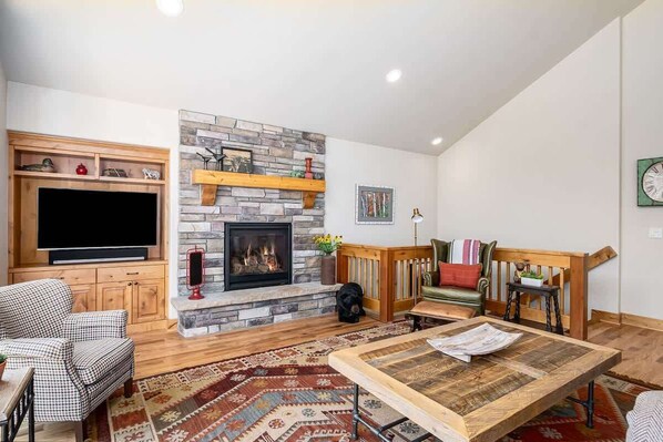Un salon avec une cheminée en pierre, des meubles en bois, un téléviseur et une horloge au mur.