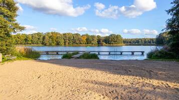 A sandy beach with a wooden dock extending into a lake, surrounded by trees.