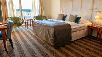 A hotel room with a bed, two chairs, a small table, a lamp, and a view through a window.