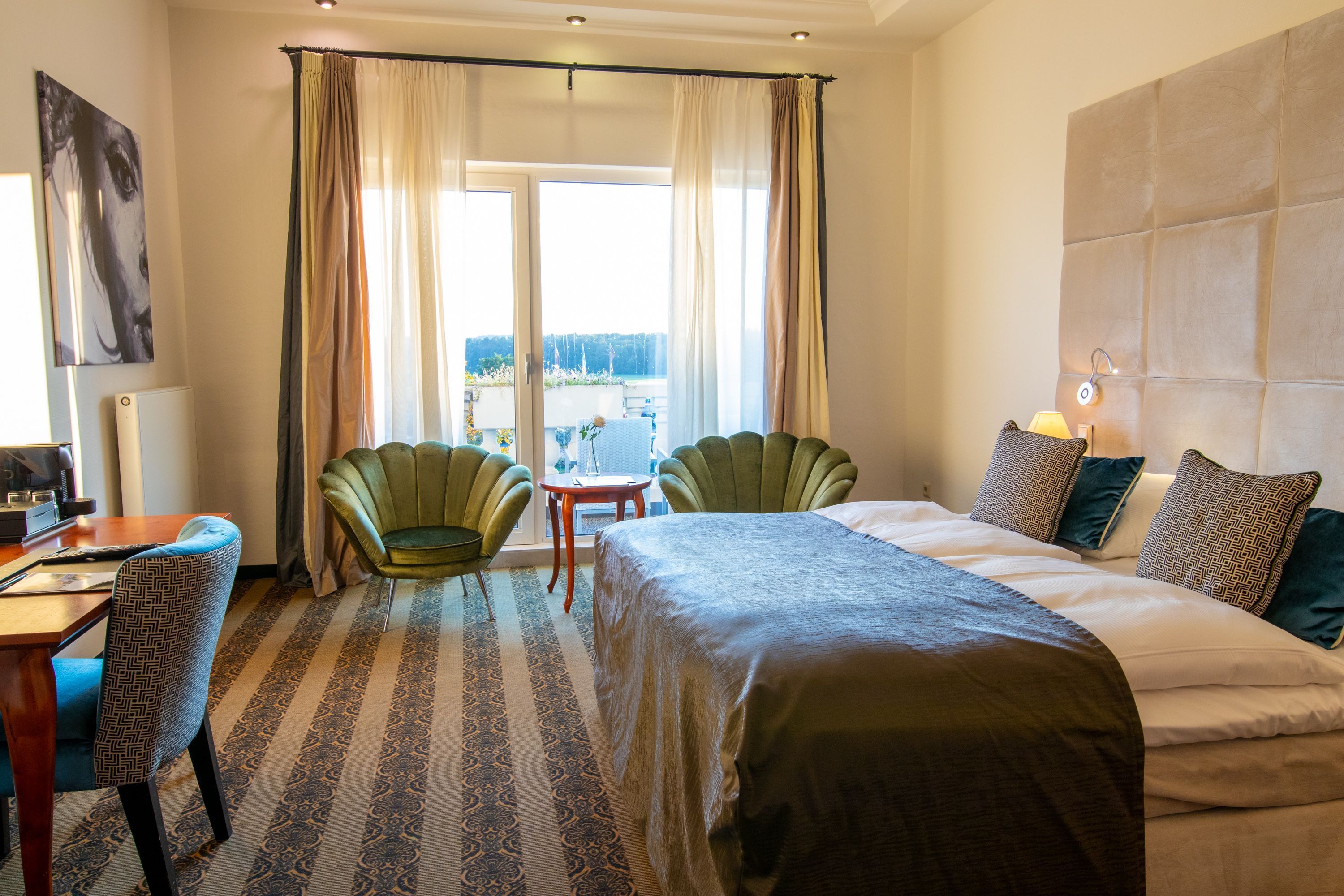 A hotel room with a bed, two chairs, a desk, a view through a window, and a headboard.