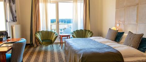 A hotel room with a bed, two chairs, a desk, a view through a window, and a headboard.