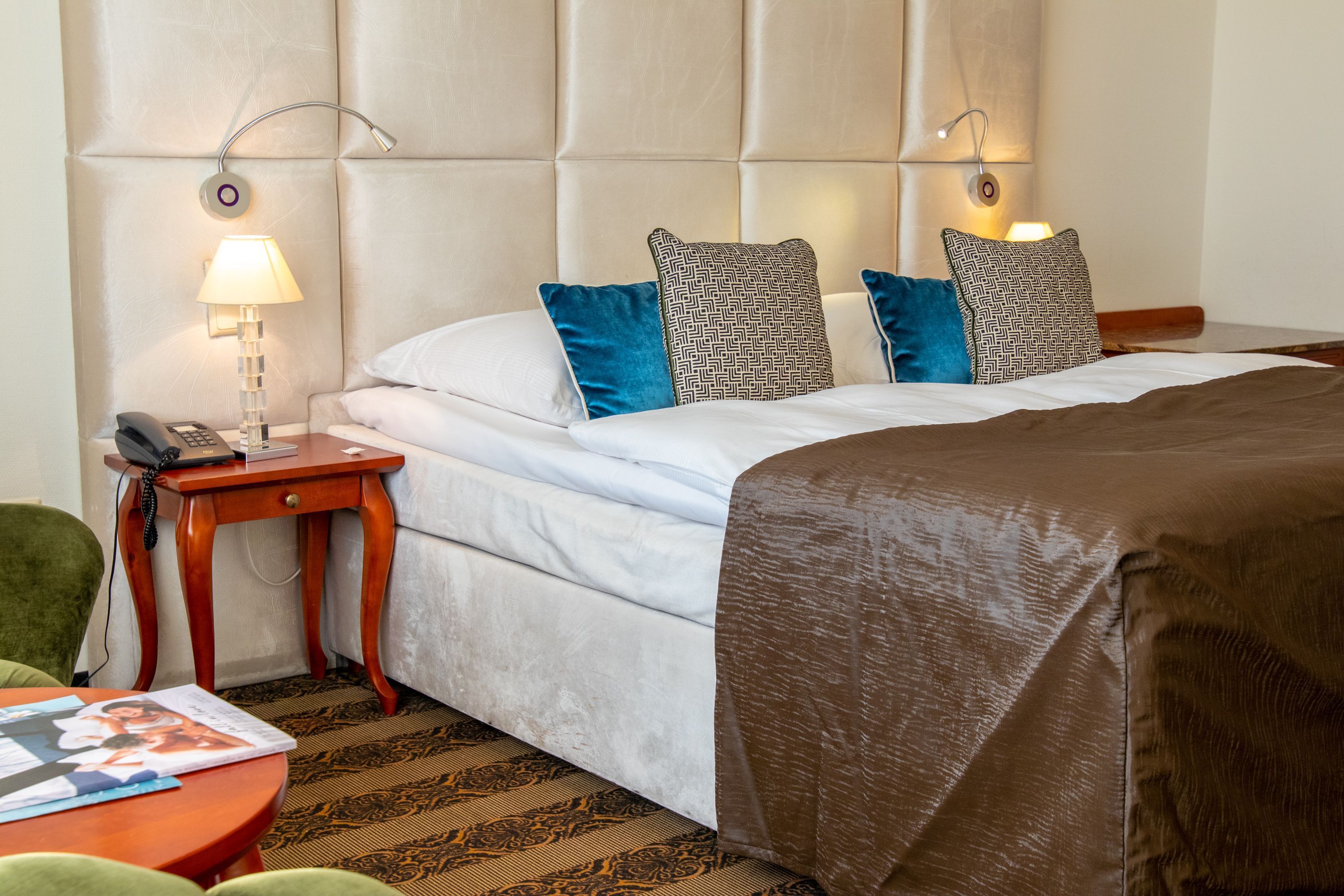 A hotel room with a bed, a bedside table with a lamp, a telephone, and a green chair.