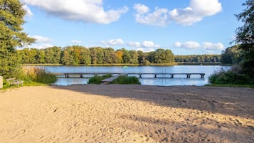 A sandy beach with a wooden dock extending into a lake, surrounded by trees.