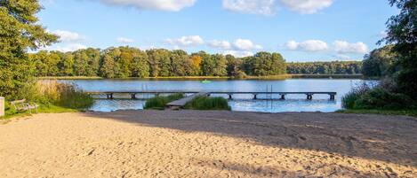 A sandy beach with a wooden dock extending into a lake, surrounded by trees.