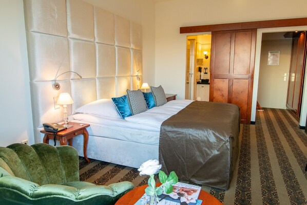 A hotel room with a bed, a green armchair, a small table with a vase of flowers, and a wooden door.