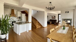 Une cuisine spacieuse avec des meubles blancs, un escalier en bois, un coin repas avec une table et des chaises en bois, et un salon doté d’une cheminée.