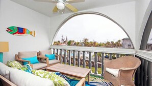Een balkon met een gebogen hekwerk, een plafondventilator en uitzicht op palmbomen en huizen.