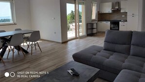 Zona d'estar espaiosa amb un sofà gris, una taula de dinar amb caires, i una cuina al fons.