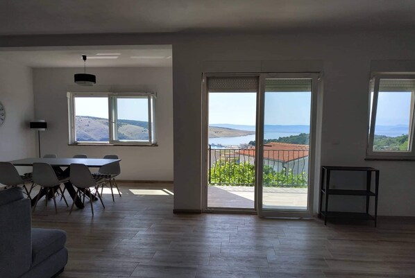 A spacious living room with a dining area, large windows, and a balcony with a view of the sea and hills.
