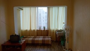 Una stanza piccola con un letto, una scrivania con un computer, una finestra con tende, una ventola a piantana e una pianta in vaso.