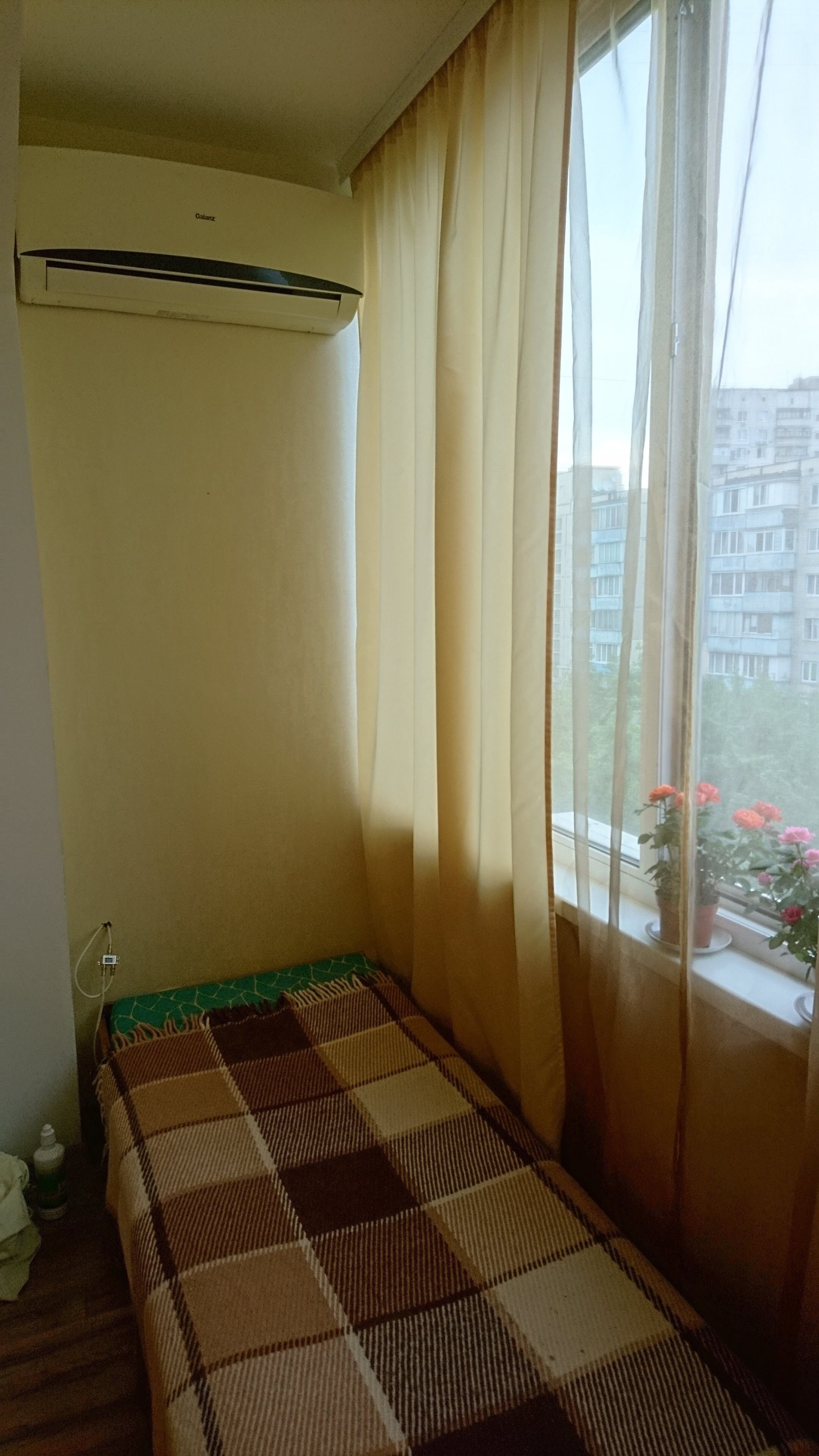 Une chambre avec un lit, un climatiseur, une fenêtre avec des rideaux et une vue sur les bâtiments et les fleurs à l’extérieur.