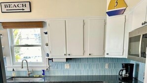 A kitchen with white cabinets, a blue backsplash, a stainless steel sink, and a trash can. - Classic Beach Cottage In Town (Rehoboth Beach)