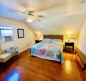 A bedroom with a wooden floor, a ceiling fan, a bed with a colorful quilt, two bedside tables with lamps, a chair, and a window with blinds. - Classic Beach Cottage In Town (Rehoboth Beach)